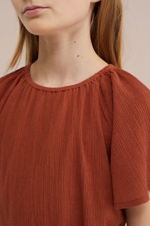 Közeli kép egy világos bőrű, vöröses hajú személyről, aki texturált, rozsda színű blúzt visel, összehúzott nyakkivágással és rövid ujjakkal.