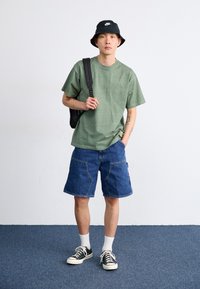Grønn bomulls-T-skjorte, blå denim cargo-shorts, svart bøttehatt, svart ryggsekk og svarte joggesko med hvite såler og hvite ankelsokker.