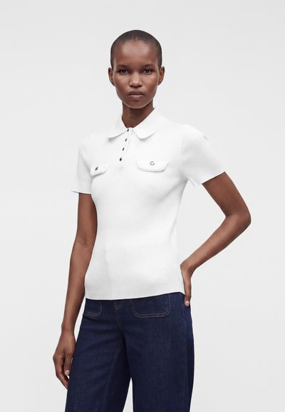 Femme aux cheveux courts porte un polo blanc côtelé à manches courtes avec des boutons et un jean bleu foncé sur un fond uni.
