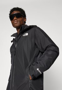 Crna vodootporna jakna s podesivom kapuljačom, prednjim zatvaračem i logom na prsima. Sadrži GORE-TEX® oznaku na rukavu. Glatka tekstura.