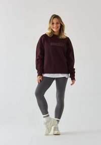 Burgundowa bluza z napisem "Księżniczka Pilates" w połączeniu z szarymi legginsami. Biała warstwowa koszula i trampki z czarno-białymi skarpetkami w paski.