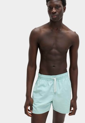 Jeune homme à la peau foncée portant un short de bain bleu clair, debout devant un fond blanc uni, faisant face à l'avant.