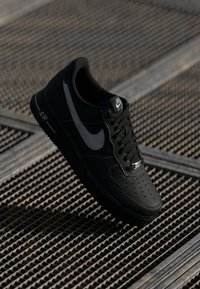 Black Nike sneaker features a textured leather upper, perforated toe, gray accents, and black laces, displayed against a gridded surface.