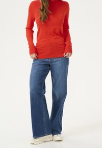 Suéter rojo de punto con cuello alto combinado con vaqueros azules de pierna ancha. El suéter tiene puños ajustados; los vaqueros son holgados con un diseño clásico de cinco bolsillos.