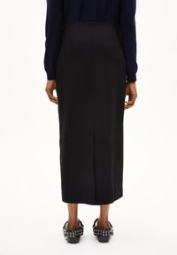 Zwarte midi-rok van glad materiaal, met een rugsplit. Gecombineerd met zwarte schoenen versierd met zilveren oogjes.