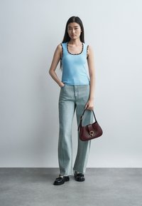 Νεαρή γυναίκα που φοράει μπλε τοπ χωρίς μανίκια και φαρδιά γκρι τζιν, κρατώντας μπορντό τσάντα, στέκει σε γκρι δάπεδο μπροστά σε φωτεινό τοίχο.