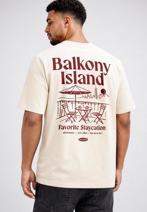 Mann mit kurzen lockigen Haaren, der ein cremefarbenes T-Shirt mit rotem Grafikmotiv einer Balkonszene und dem Text „Balkony Island Favorite Staycation“ trägt.