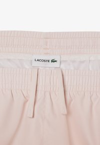 Shorts rosa chiaro con vita elastica, lacci regolabili e fodera in rete bianca. Presentano un'etichetta del logo Lacoste.
