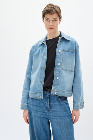 Giacca di jeans blu chiaro con colletto, tasca frontale e chiusure a bottone argentate. Abbinata a pantaloni di jeans blu scuro.