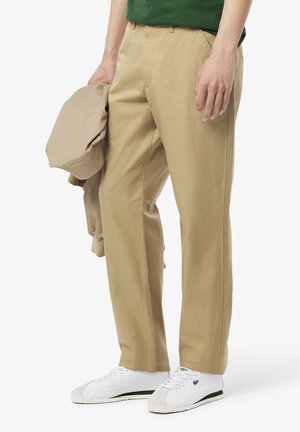 Chinos beige de algodón, con un diseño de pierna recta, bolsillos laterales y pasadores para cinturón, combinados con una camiseta verde y zapatillas blancas.