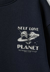 Sötét tengerészkék pulóver texturált mintával, amelyen egy szív és egy bolygó grafika látható, a "SELF LOVE PLANET" felirattal fehér, kopottas stílusban.