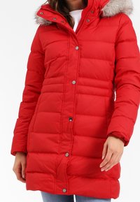 Röd puffig jacka med huva och fuskpäls på kanten. Jackan har ett quiltat mönster, tryckknappar och två sidofickor.