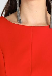 Czerwona sukienka z szerokim, zaokrąglonym dekoltem. Posiada gładką teksturę, centralny szew z przodu oraz metalowe kolczyki z frędzlami.