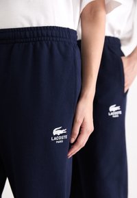 Tumši zili sporta bikses ar elastīgu jostu un sānu kabatām; kreisajā augšstilbā ir baltais Lacoste logo un teksts. Gluda tekstūra.
