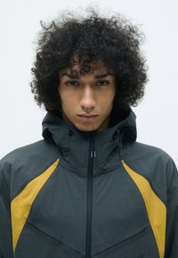 Hooded jacket tamsiai pilkos spalvos su geltonais akcentais, turinti užtrauktuką ir sportinį dizainą, pagaminta iš lygaus, lengvo audinio.