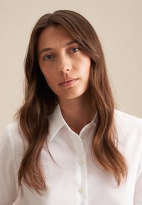 Camicia bianca con colletto classico, texture liscia e design aderente, arricchita da una sottile lucentezza. Lunghi capelli castani e ondulati.