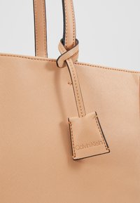 Perzikkleurige tote bag met een gestructureerd oppervlak, zwarte biezen en een hangtag met het logo. Dubbele handvatten met versterkte stiksels.