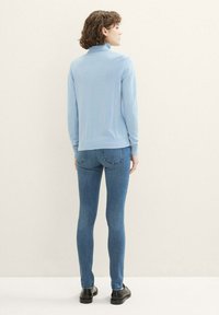 Top de manga larga azul claro con un cuello alto, combinado con jeans ajustados azules y zapatos negros, visto desde atrás. Fondo liso.
