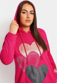 Vaaleanpunainen huppari, jossa on edessä kolmella sydämellä varustettu kuvio, joka sisältää hopeista kimallusta, punaisia pisteitä ja mustaa, tekstuurimaista kangasta. Mukana kiristettävä huppu.