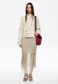 Maglione beige a maglia con collo alto, indossato su una gonna beige strutturata con pannelli trasparenti, abbinato a una borsa rossa e sandali neri.