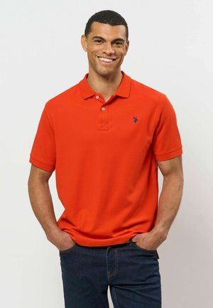 Homme souriant portant un polo à manches courtes orange vif et un jean bleu foncé, les mains dans les poches, sur un fond uni.