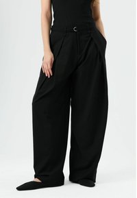 Pantalon noir taille haute à plis, jambes larges, avec passants de ceinture et boucle circulaire argentée, fabriqué en tissu lisse.