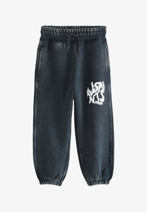 Persona che indossa pantaloni della tuta neri con scritta bianca "AllSaints London" sulla coscia, abbinati a sneakers bianche e una felpa con cappuccio nera.