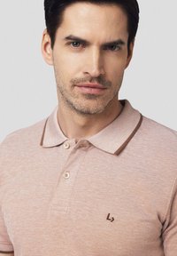Jasnoróżowa koszulka polo z teksturowanym wzorem, kontrastującym brązowym kołnierzykiem i trzema guzikami. Na piersi znajduje się małe brązowe logo.