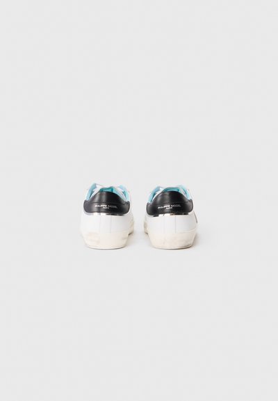 Baskets blanches avec des accents noirs au niveau du talon et des lacets bleus. L'arrière présente un logo et une semelle légèrement usée, indiquant une utilisation.