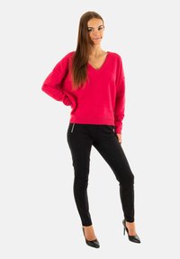 Felroze oversized sweater met een V-hals, gecombineerd met zwarte slim-fit broek en zwarte hoge hakken. Gladde textuur en casual ontwerp.