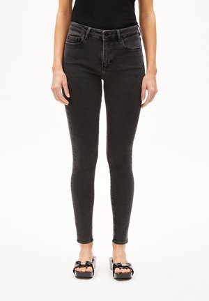 Zwarte slim-fit jeans met een hoge taille, voorzakken en riemlussen. De stof heeft een gladde textuur met een lichte stretch. Gemaakt in combinatie met zwarte plateau-sandalen.