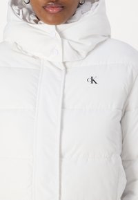 Biely puffer kabát s vysokým golierom, so značkou na ľavej strane a cvočkami, vyrobený z hladkého, textúrovaného materiálu.