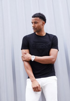 Muž v čiernom tričku Guess a bielych nohaviciach stojí pri šedej stene, pozerá doľava s prekríženými rukami a nosí biele hodinky.