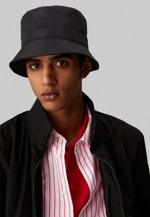Chapeau seau noir en tissu lisse, avec une étiquette sur le devant. Porté avec une veste noire sur un polo rouge et une chemise rayée.