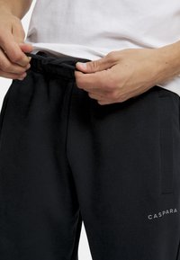 Svarte joggebukser med elastisk liv, snøring, sidelommer, myk tekstur og kontrasterende "CASPARA"-logo på nederste del av benet.