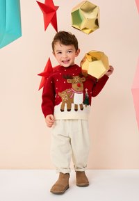 Pull en tricot rouge avec motif de renne, bas crème, et petits bottillons marron. Pantalon en velours côtelé blanc, décorations en papier colorées en arrière-plan.