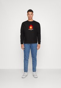 Czarna bluza z haftowanym wzorem ogniska, zestawiona z niebieskimi dżinsami i białymi sneakersami. Model stoi na tle jednolitej ściany.