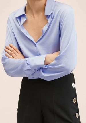 Personne portant une chemise bleu clair à manches longues avec col et un pantalon noir avec une rangée de gros boutons sur le côté, les bras croisés.