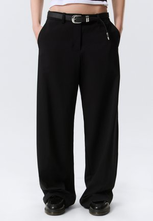 Person som har på seg løse svarte bukser med svart belte og svarte sko, hendene i lommene, står foran en ensfarget hvit bakgrunn.