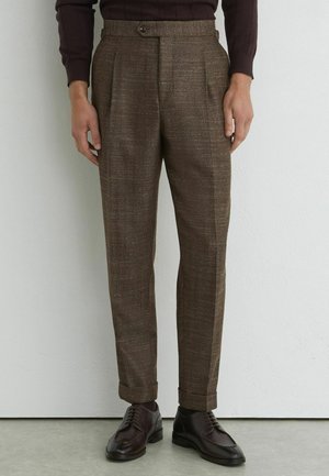Man draagt bruine herringbone op maat gemaakte broek met omslagen en donkerbruine nette schoenen, staand tegen een witte muur.