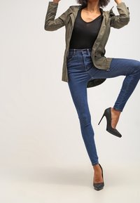 Olivově zelená bunda, černý přiléhavý top, modré skinny džíny a černé vysoké podpatky. Bunda má zipový uzávěr a kapuci.