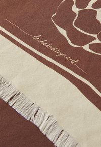 Barna szövet, fehér grafikai dizájnnal, rojtos szélekkel és nyomtatott szöveges részlettel. Simább textúra, kontrasztos színekkel.