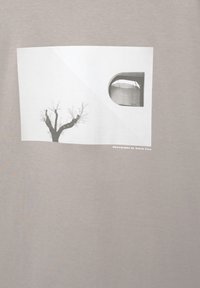 Grå T-shirt med et hvidt grafisk tryk af et bart træ og et vindue, trykt på et glat bomuldsstof.