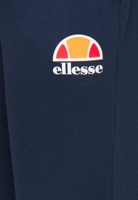 Das Ellesse-Logo auf den navyblauen Sweatpants zeigt einen bunten Bogen in Orange, Gelb und Rot. Weicher Stoff mit glatter Textur.