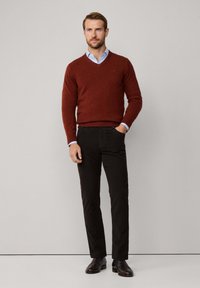 Brauner V-Ausschnitt-Pullover aus weichem Strick; hellblaues Hemd mit Kragen darunter; dunkle Hose; glänzende Schuhe, Hände in den Taschen, neutraler Hintergrund.