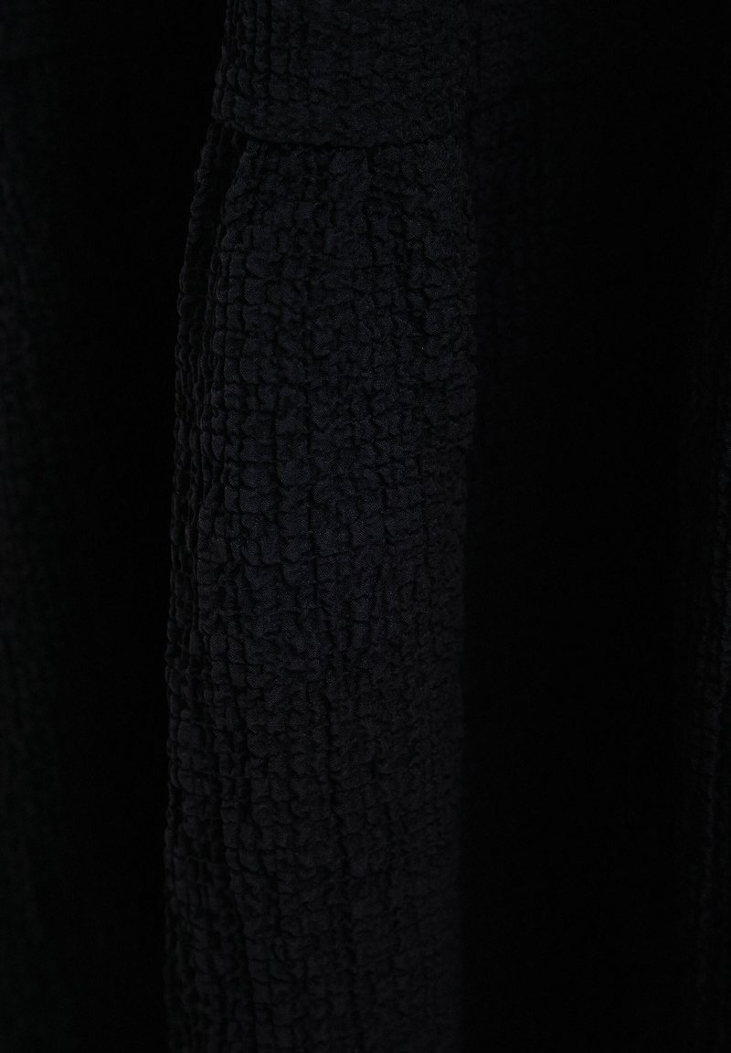 Czarna fakturowana tkanina z pofalowaną powierzchnią, z fałdami i głębokością, tworząca subtelny wzór. Brak widocznych elementów metalowych lub akcentów.