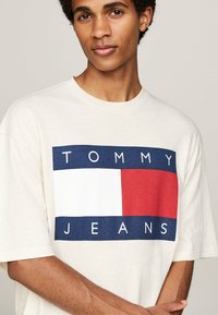 Camiseta color crema con un gran gráfico en navy, rojo y blanco que presenta el texto "TOMMY JEANS" en letras de bloque. Mangas cortas y corte relajado.