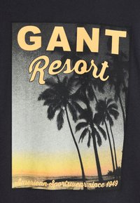 Camiseta gráfica negra con la silueta de una palmera contra un atardecer, con "GANT Resort" y "American Sportswear desde 1949" en amarillo.