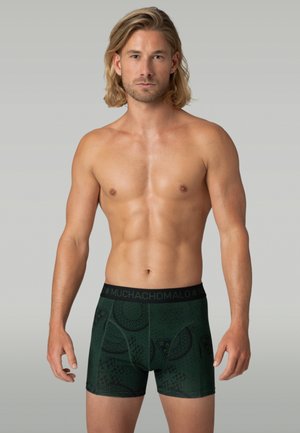 Muž bez trička s blond vlasmi po plecia, oblečený v tmavozelených boxerkách s motívom a pásom s nápisom "Muchachomalo", stojí na sivom pozadí.