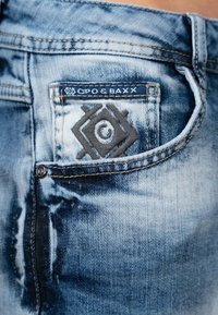 Jean en denim avec un délavage bleu, des coutures contrastantes, une patch sur la poche arrière et une étiquette de logo sur la ceinture.